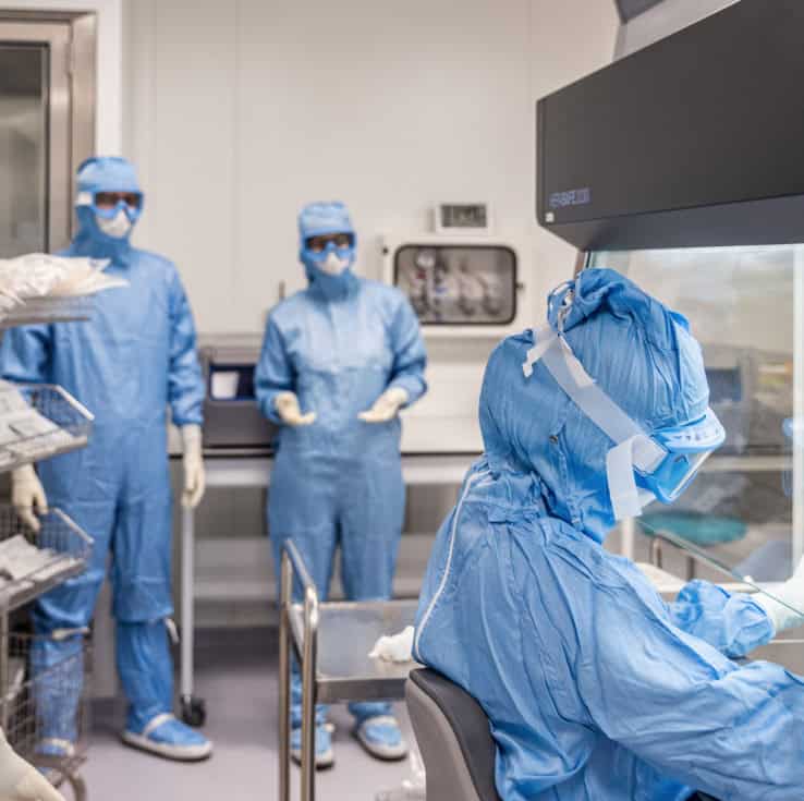 Four technicians working in a GMP manufacturing environment. One is performing manipulations inside a biosafety cabinet performing manipulations, while the other three stand in the background.- Cell-Easy