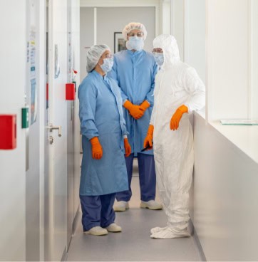 Three employees in cleanroom attire talking in a modern, sterile cleanroom corridor. Colored signal lights are visible on the left wall. – Cell-Easy